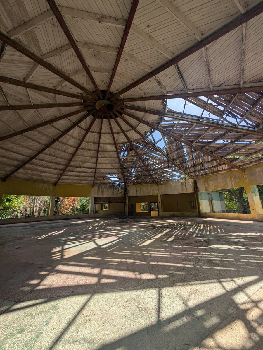 Inside the old octagonal church: timber roof beams radiate from the centre, part of the roof is open to the sky, and sunlight falls across the floor